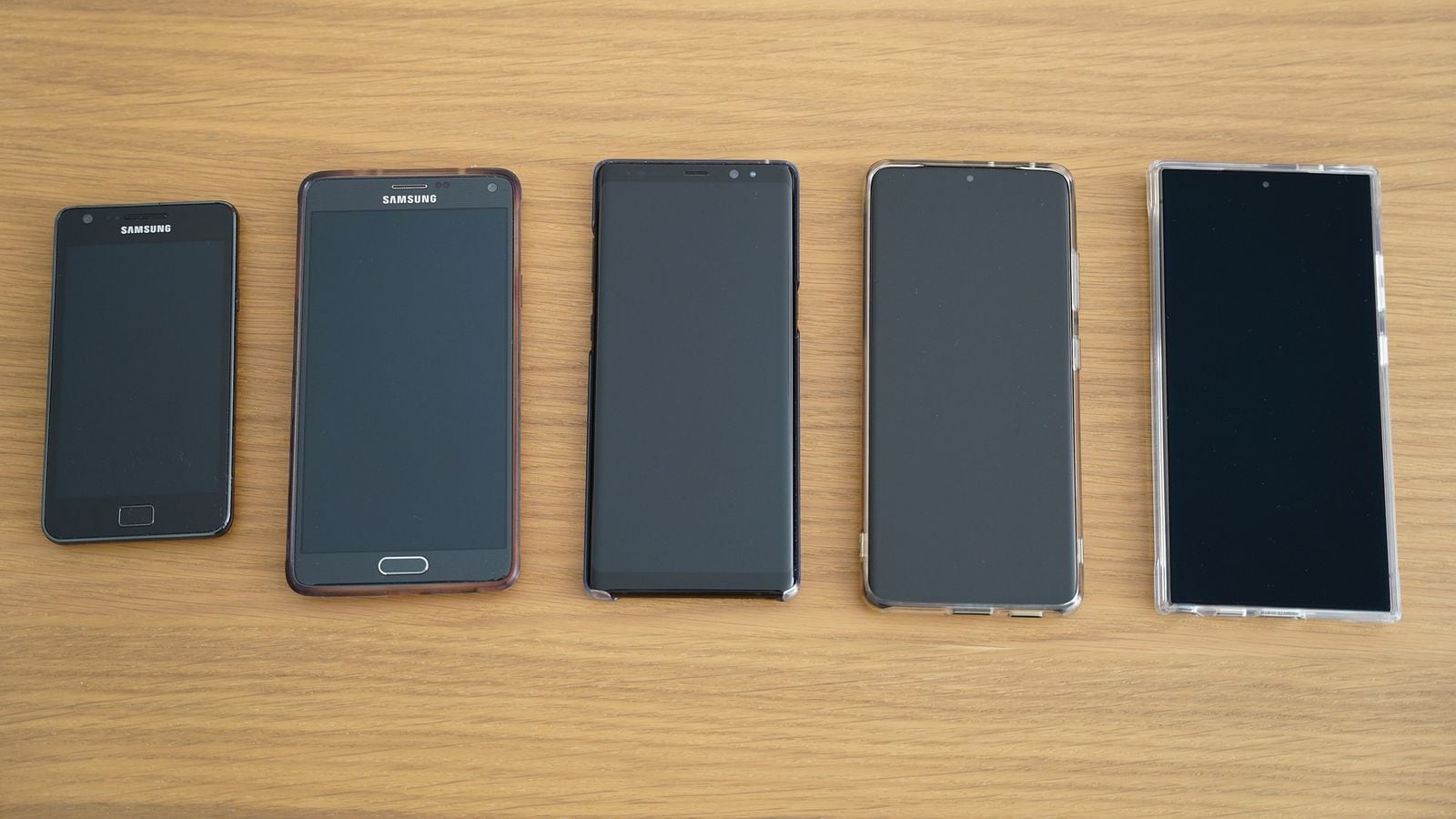 Pět telefonů Samsung Galaxy (S2, Note4, Note8, S21, Ultra v pouzdru) vyrovnaných na dřevěném stole